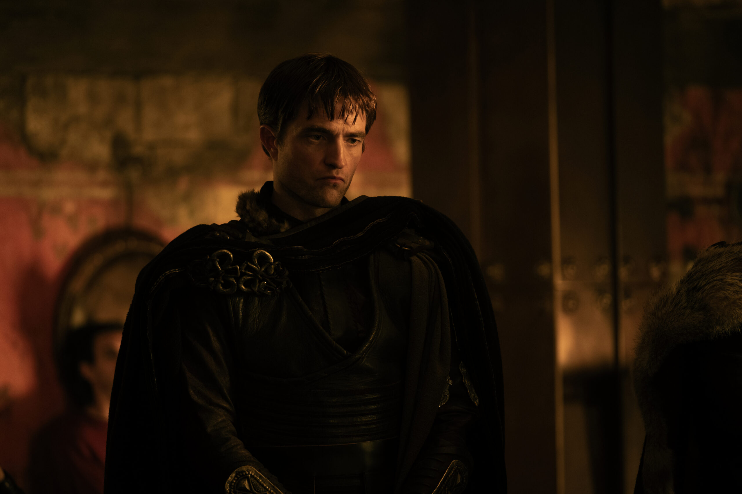 Dark, amber-lit scene of a man in heavy medieval armor looking down pensively in a dim hall.