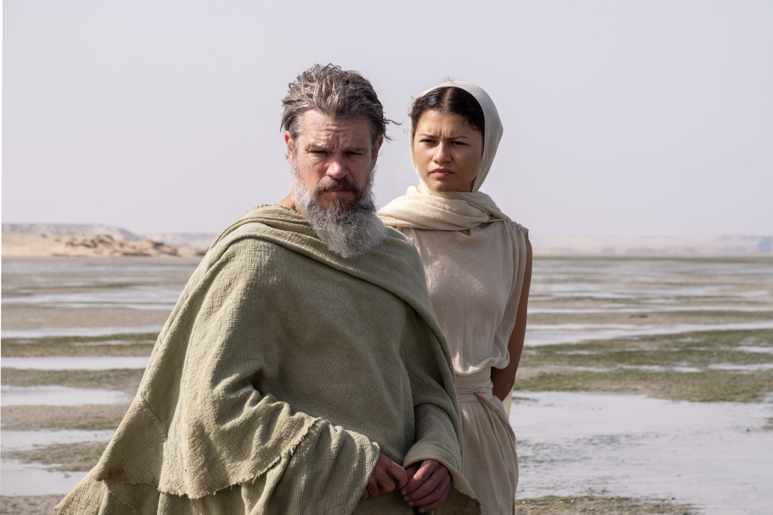 Two people in beige robes stand together on a desolate shoreline with shallow water and distant rocks, looking toward the camera with serious expressions.