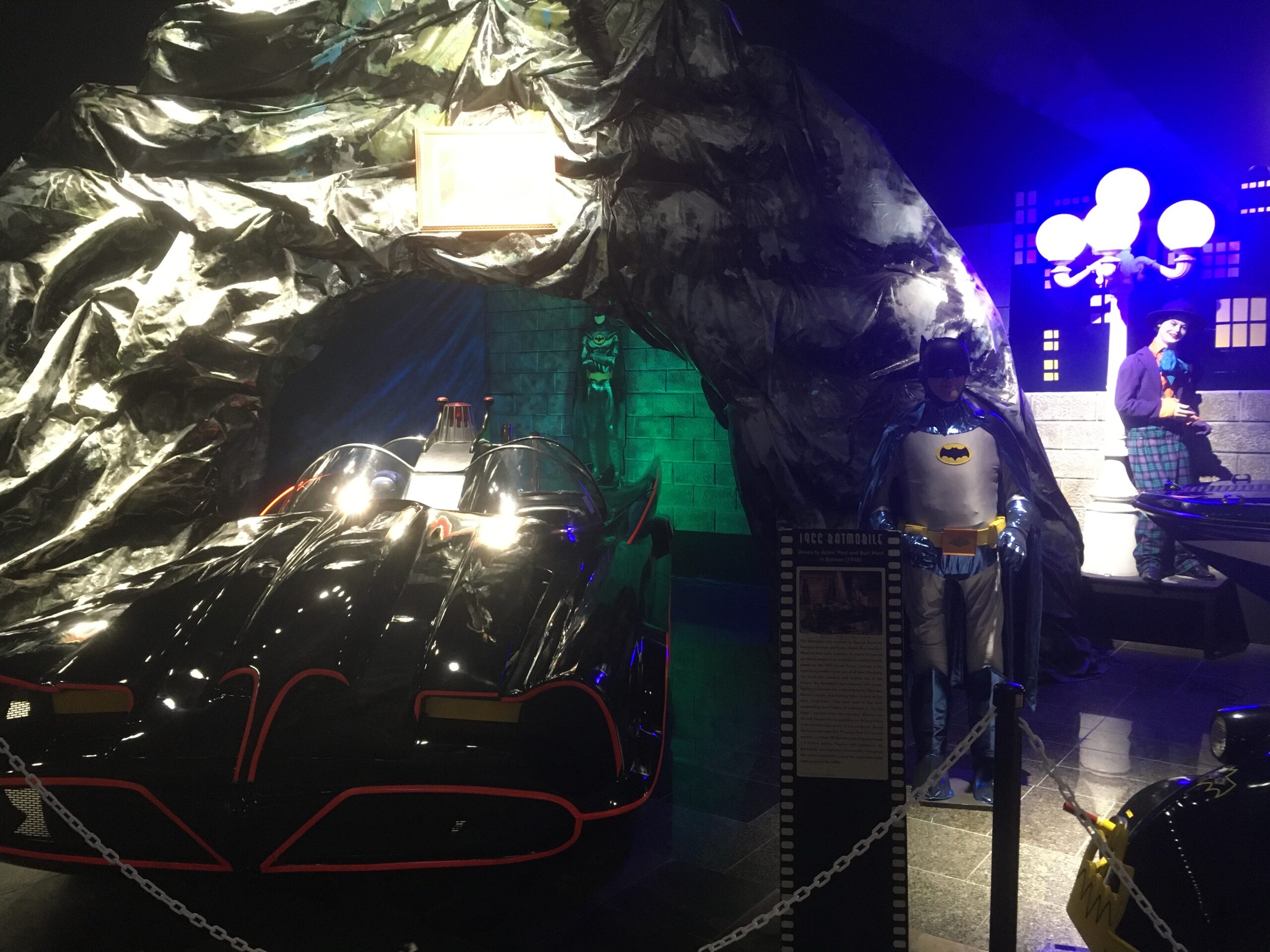 A black Batman-era car on display, with red trim, under dark, crinkled tarps in a museum setting and a Batman figure nearby in costume.